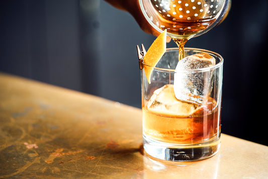 Ein Glas Whiskey mit einer Orangenzeste und Eis wird durch ein Sieb aus einem Shaker eingegossen | Old Soggy Spiced Bourbon